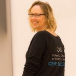 Middle aged white, blonde woman in Theatretrain top with Cas written on it, looking back at the camera