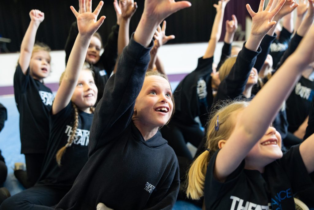 Child smiling during a performing arts class while trying a new activity with support from other young performers.